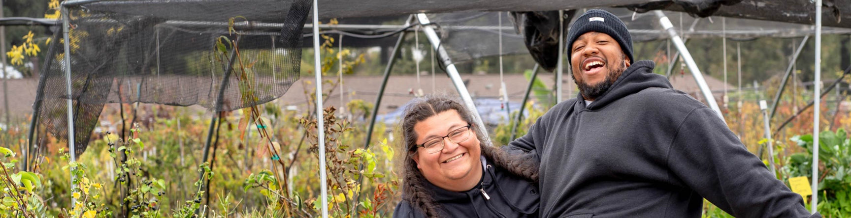 Two PJ staff members embracing each other with one leg up in front of a lush nursery background